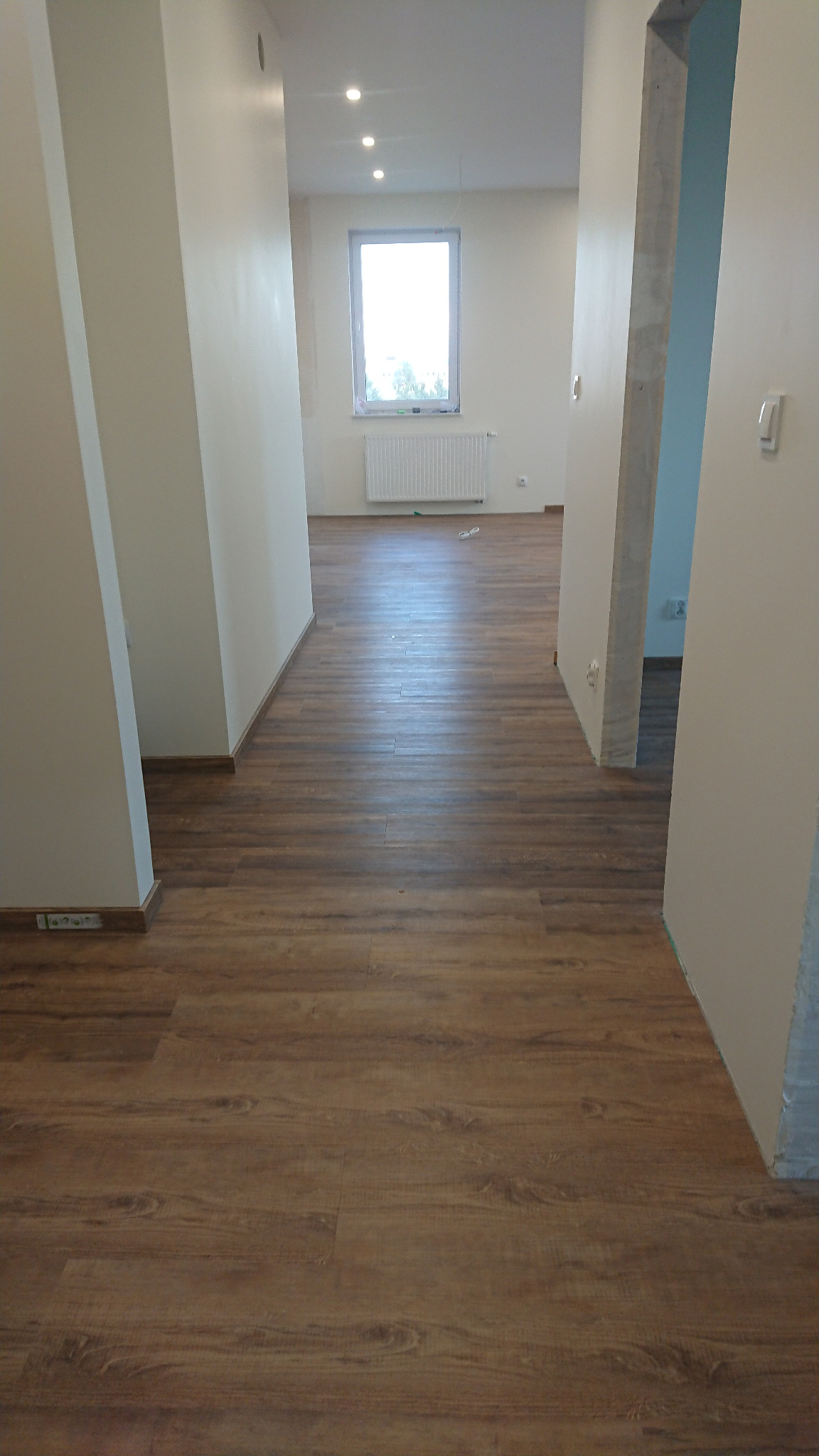 Wykończone wnętrze z drewnopodobną podłogą, świeżo pomalowanymi ścianami na biało i widocznym fragmentem surowego, betonowego filaru w przejściu.