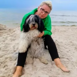 Kobieta w zielonej bluzie i okularach siedzi na piasku na plaży, trzymając psa rasy owczarek holenderski długowłosy.