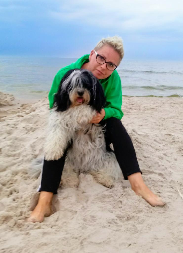 Kobieta w zielonej bluzie i okularach siedzi na piasku na plaży, trzymając psa rasy owczarek holenderski długowłosy.