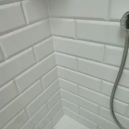 Wykończona kabina prysznicowa w Udaninie z białymi, prostokątnymi płytkami ułożonymi w cegiełkę i elementami czarnych detali na podłodze, widoczny fragment słuchawki prysznicowej.