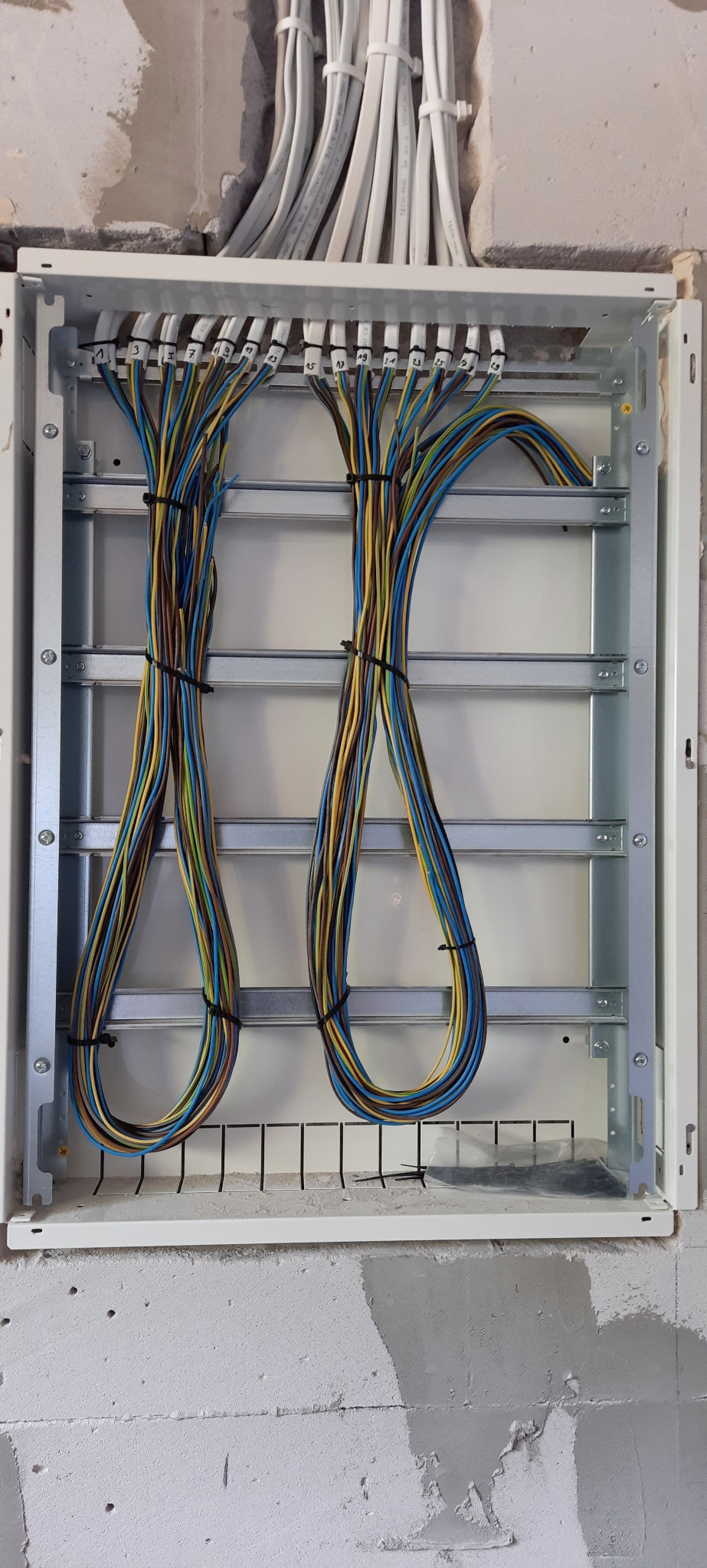 Wewnętrzny widok metalowej skrzynki rozdzielczej z uporządkowanymi kablami oznaczonymi numerami, zebranymi w wiązki za pomocą czarnych opasek zaciskowych, zamontowanej na surowej ścianie.