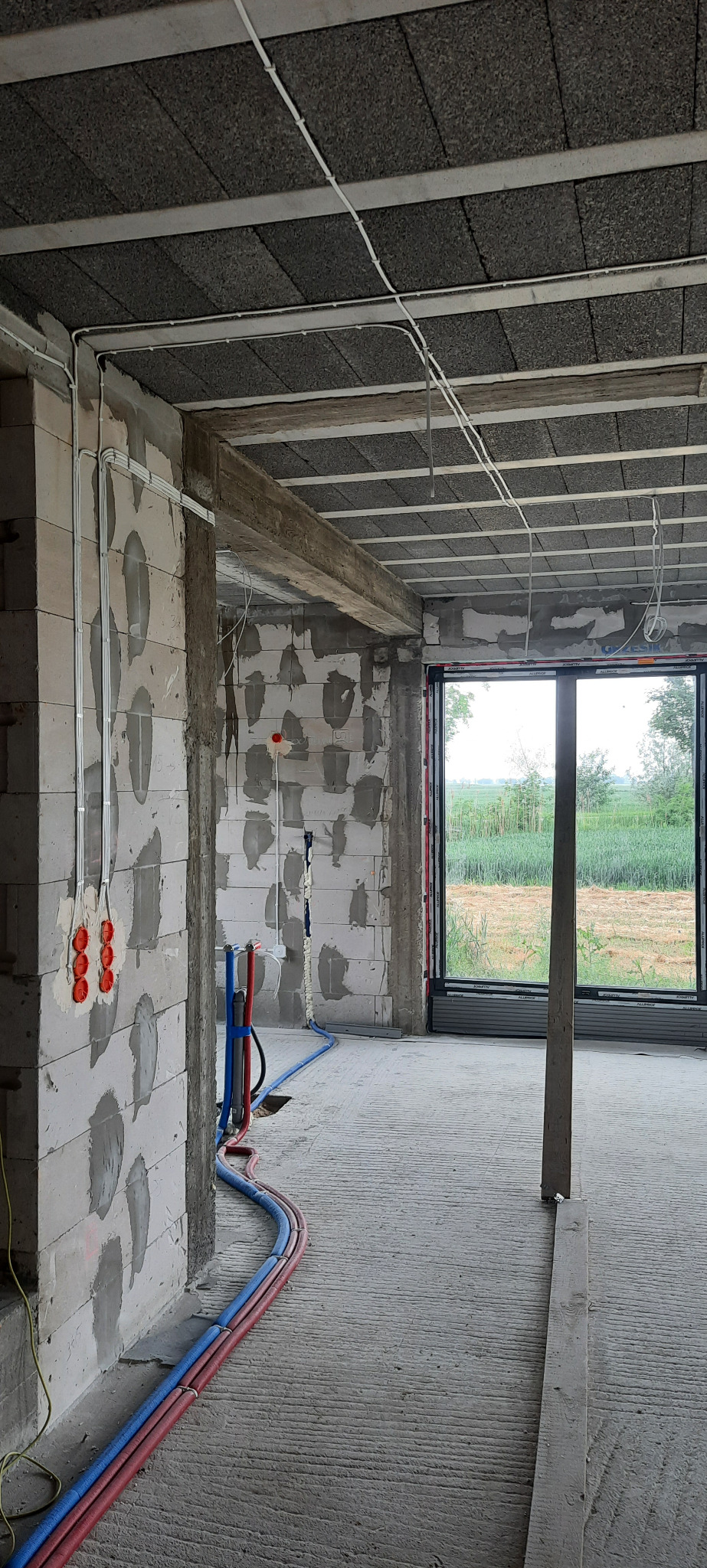 Wnętrze w surowym stanie z widocznymi przewodami elektrycznymi, rurami instalacji wodnej i gniazdkami w ścianie z betonu komórkowego, duże okno z widokiem na pole.