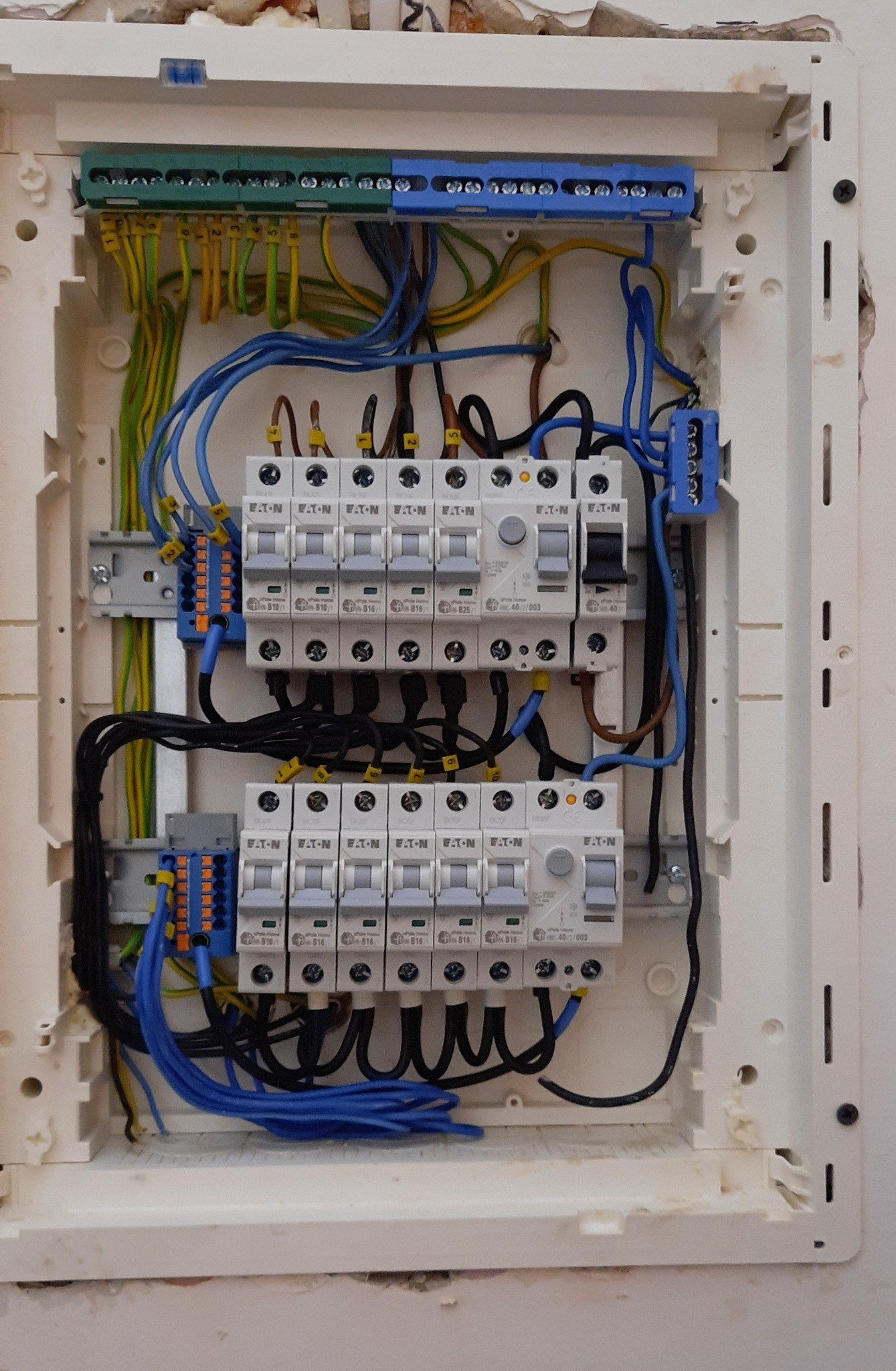 Otwarta skrzynka elektryczna z zainstalowanymi bezpiecznikami EATON, widoczne kable w różnych kolorach, listwy zaciskowe i fragment ściany z ubytkami tynku.