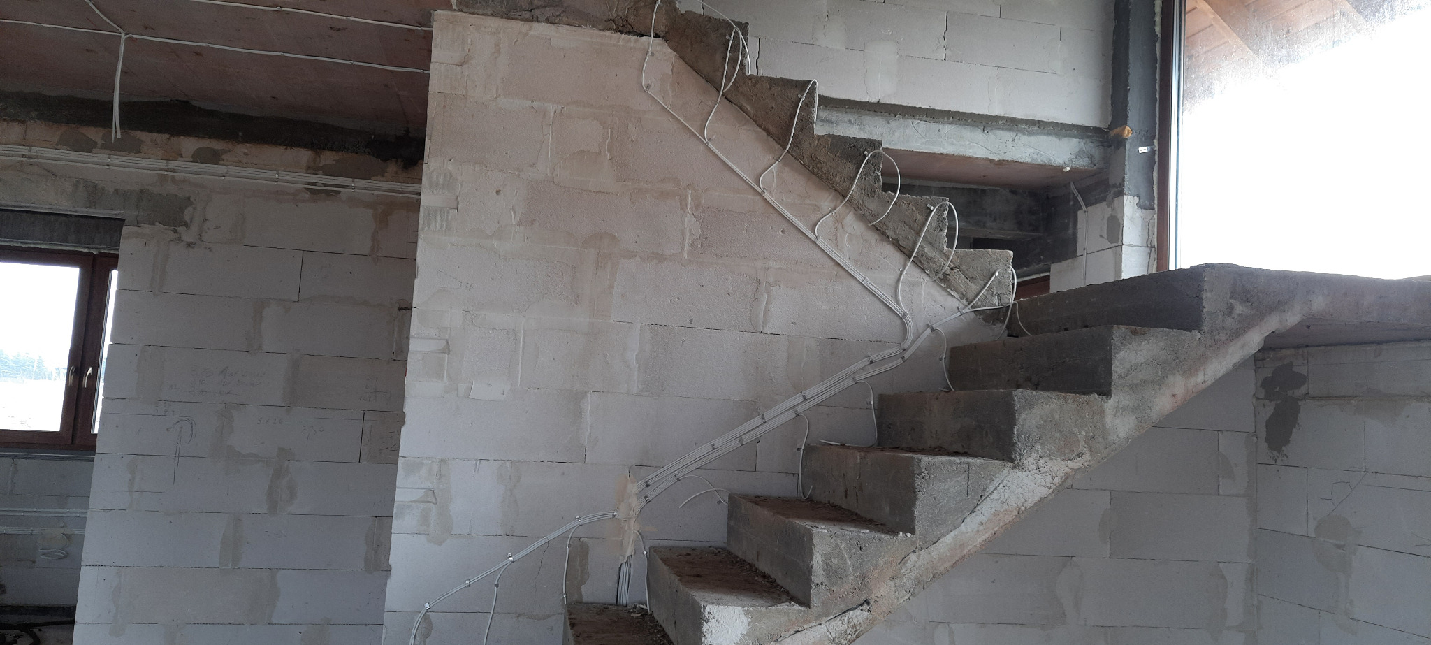 Surowe betonowe schody w trakcie budowy, z poprowadzonymi przewodami elektrycznymi wzdłuż ściany i stopni, widoczne białe ściany z betonu komórkowego.