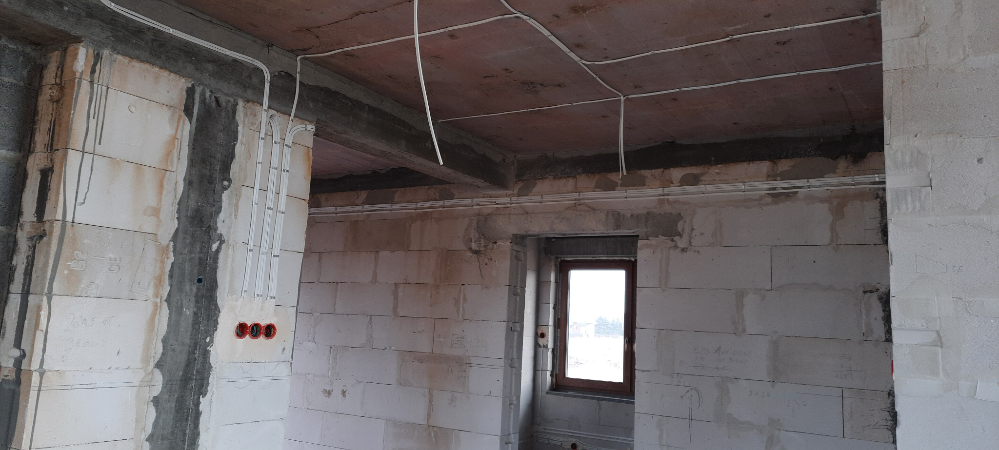 Wnętrze w stanie surowym z widoczną instalacją elektryczną: kable w białych rurkach prowadzone po ścianach i suficie, czerwone gniazdka elektryczne zamontowane w ścianie z betonu komórkowego.