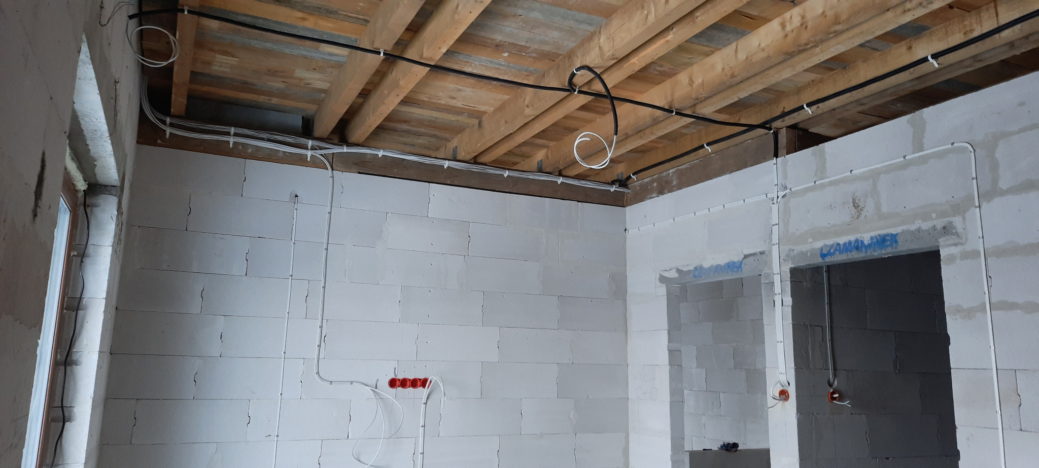 Wnętrze budynku w Nowym Stawie w trakcie prac elektrycznych: widoczne kable prowadzone po drewnianym stropie i ścianach z białych bloczków, z przygotowanymi puszkami instalacyjnymi.