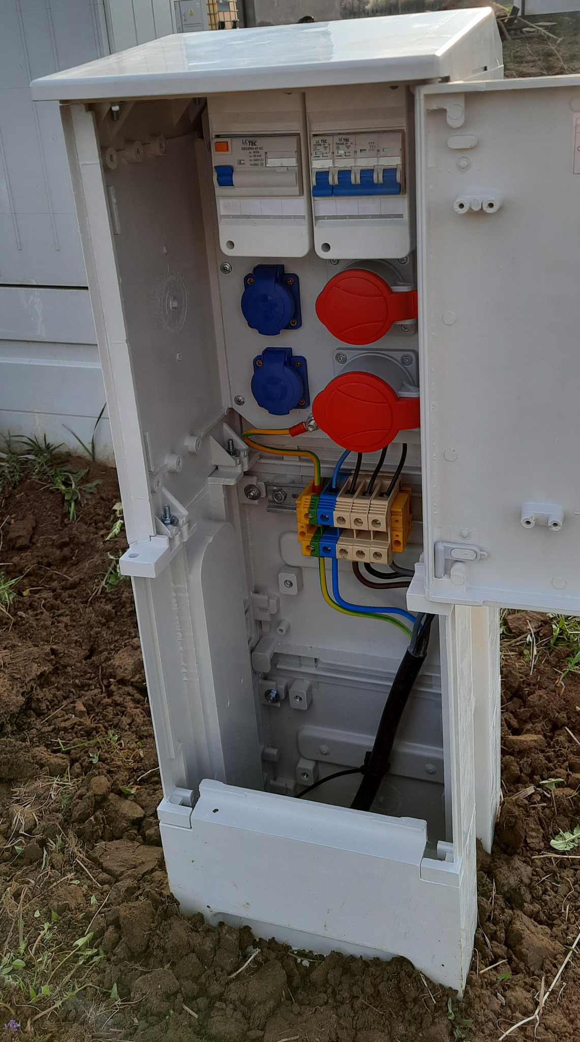 Otwarta skrzynka elektryczna z widocznymi bezpiecznikami, gniazdami przemysłowymi (czerwone i niebieskie) oraz połączeniami kablowymi, umieszczona w ziemi.