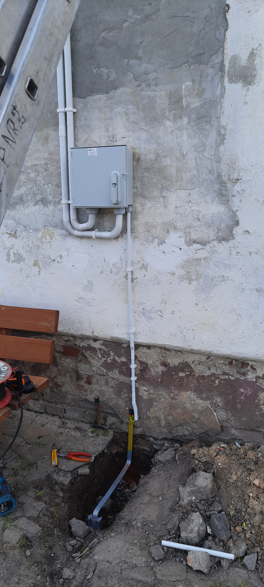 Szara skrzynka elektryczna z białymi rurami na starej, łuszczącej się ścianie budynku, widoczny wykop z kablem w ziemi i narzędzia pracy. Metalowa drabina oparta o budynek z lewej strony.