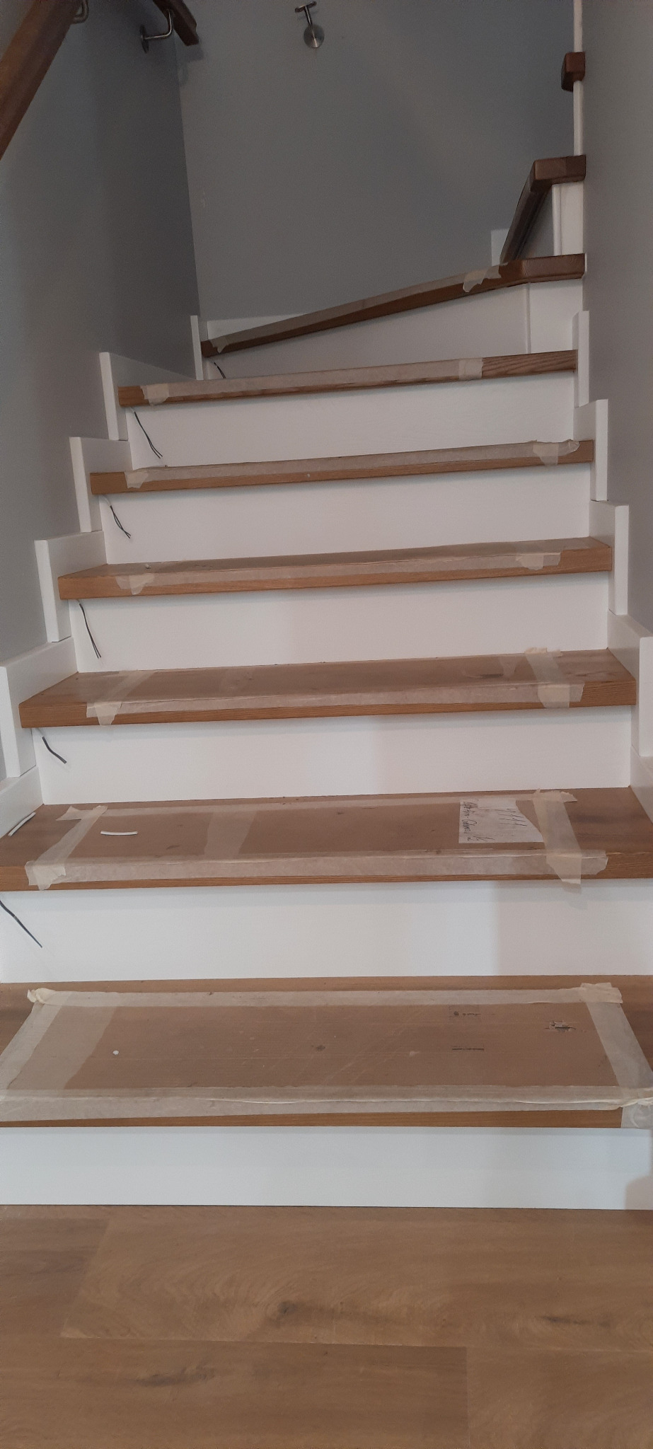 Schody drewniane z białymi podstopnicami, zabezpieczone folią malarską podczas prac wykończeniowych, widoczne przewody elektryczne wystające ze ściany.