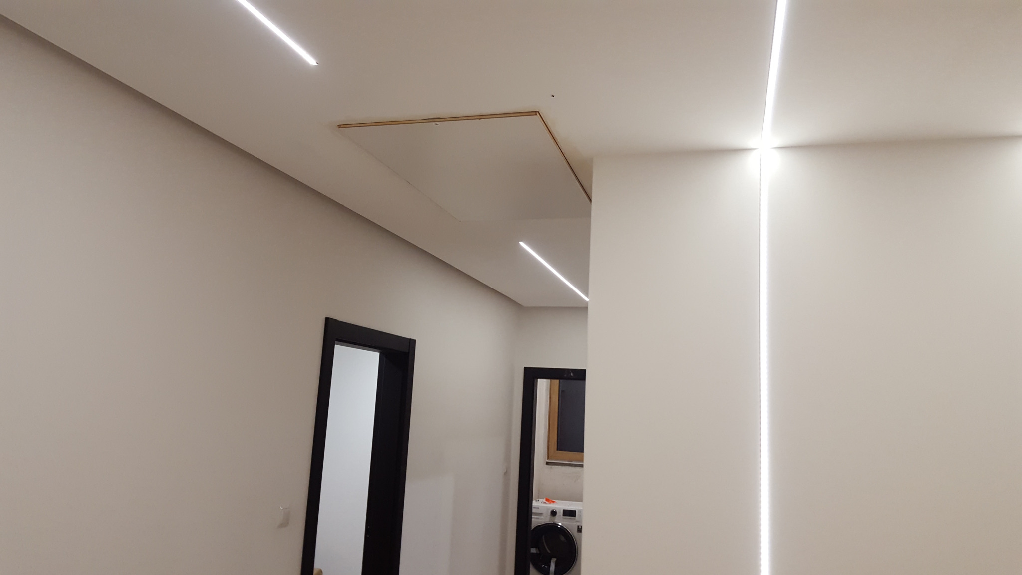 Nowoczesne oświetlenie LED liniowe w minimalistycznym wnętrzu, widoczna klapa rewizyjna na suficie, czarne ramy drzwi w tle.