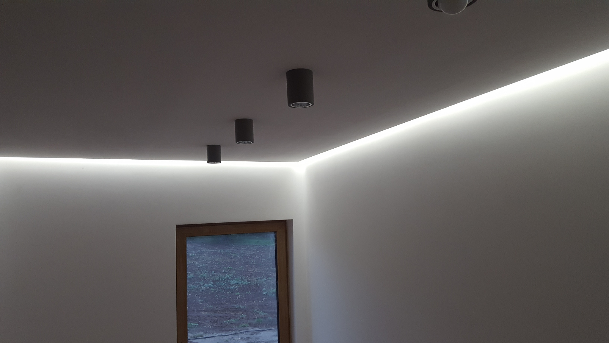 Minimalistyczne oświetlenie sufitowe: trzy czarne, cylindryczne lampy punktowe i listwa LED wzdłuż krawędzi sufitu, nad drewnianą ramą okna.