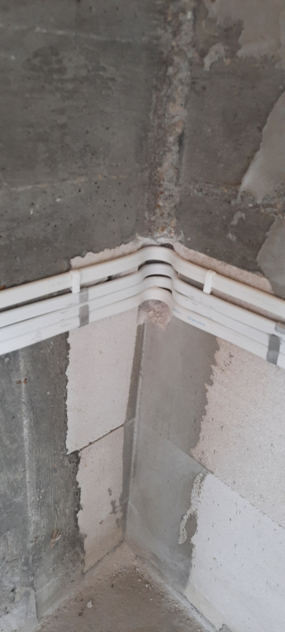 Narożnik sufitu i ścian w surowym stanie z zamontowanymi trzema białymi przewodami elektrycznymi w peszlu, przytwierdzonymi do ściany szarymi uchwytami.