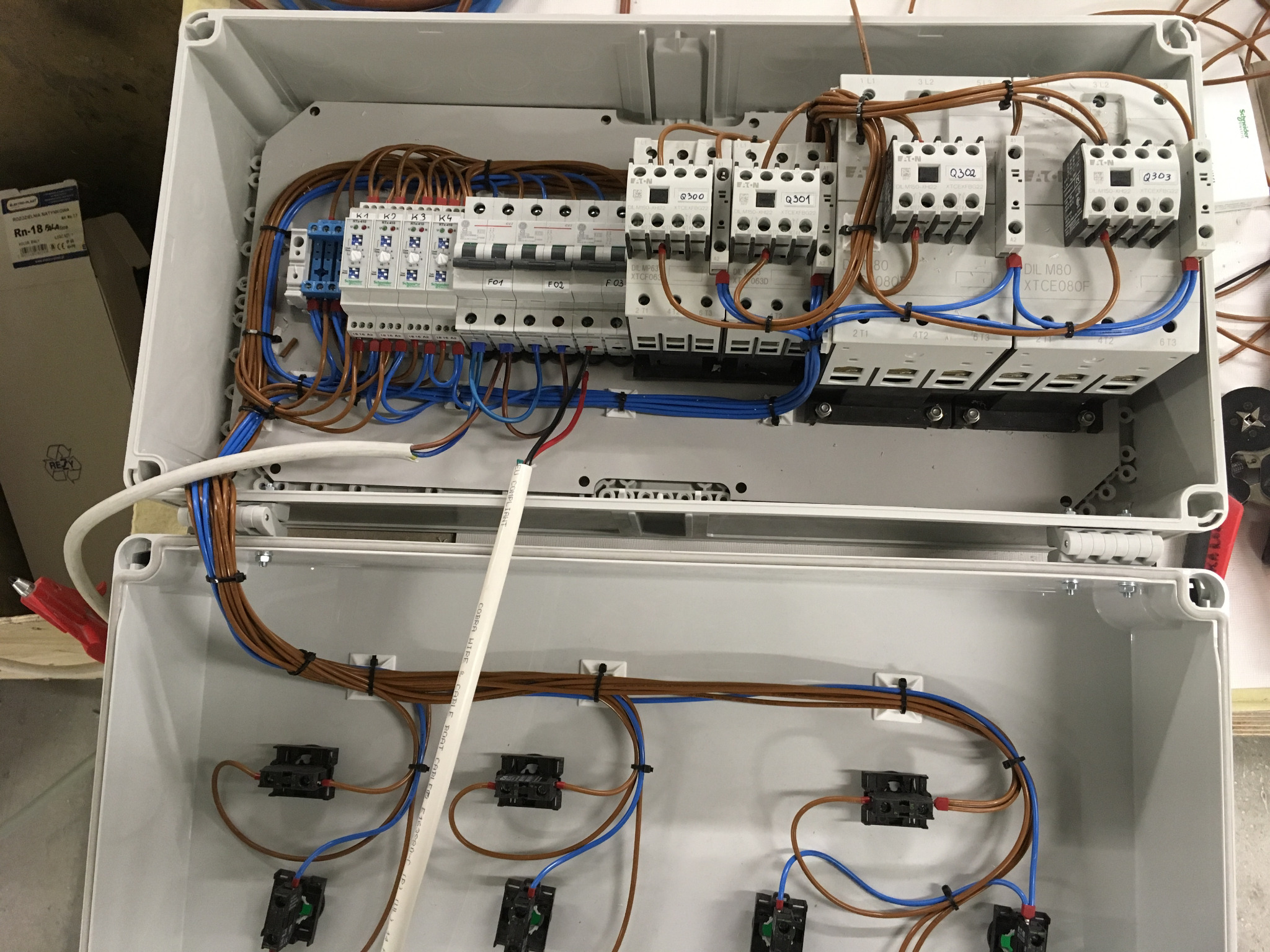 Otwarta skrzynka rozdzielcza z zamontowanymi bezpiecznikami, stycznikami i okablowaniem w kolorach brązowym i niebieskim, widoczne etykiety z oznaczeniami elementów.