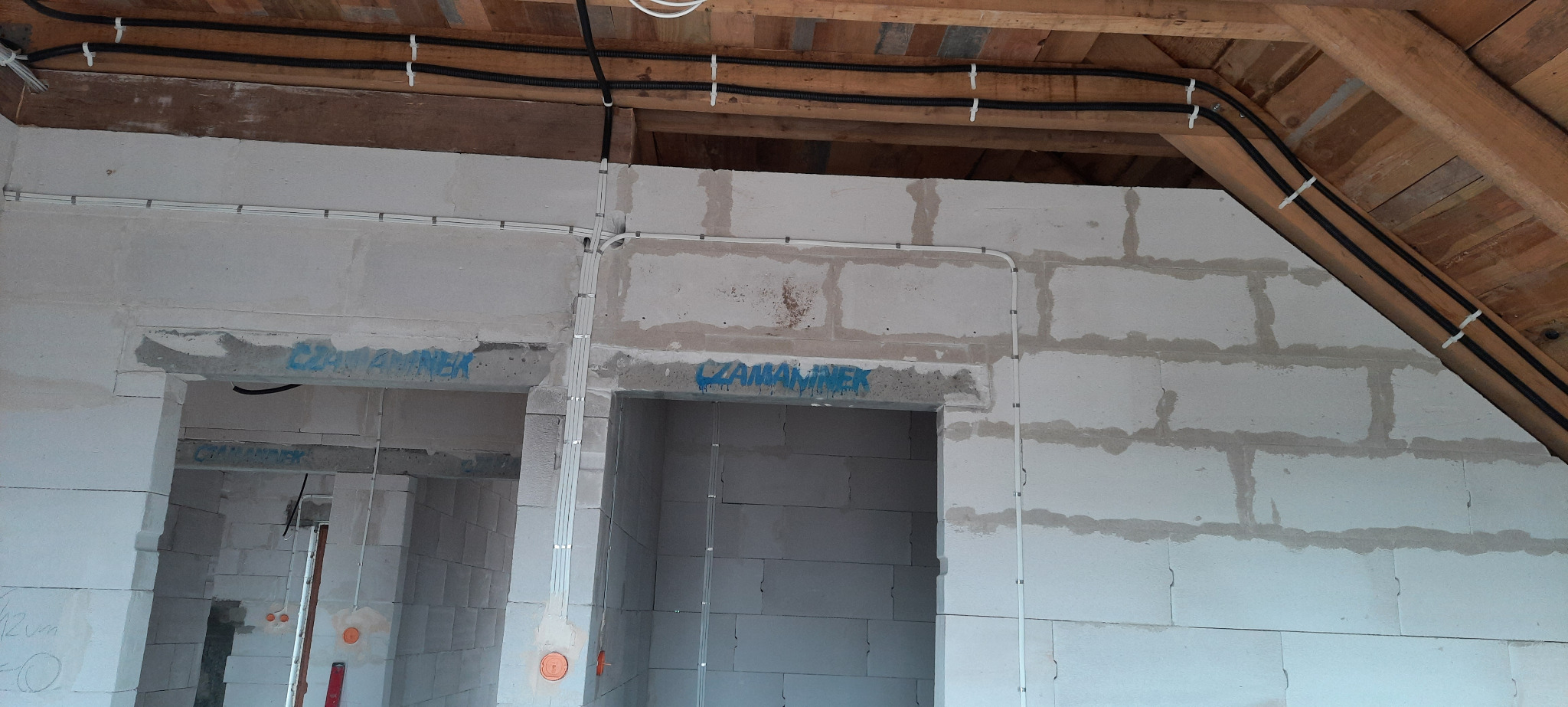 Instalacja elektryczna w surowym budynku z białych bloczków, widoczne kable prowadzone po ścianach i pod sufitem, mocowane białymi uchwytami, zarys otworów drzwiowych z napisem 'CZARNAMINEK'.