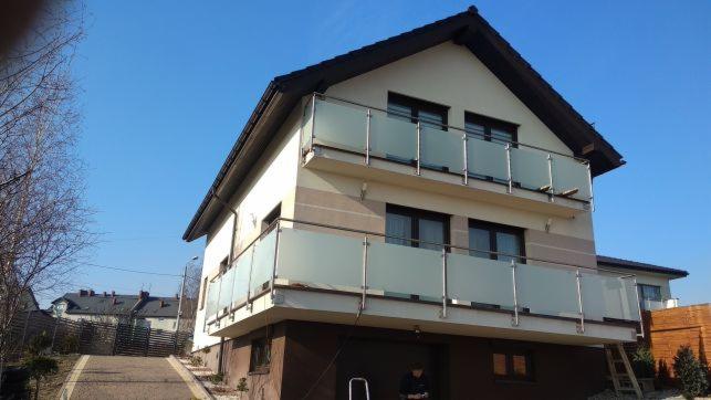 Dwukondygnacyjny dom z balkonami zabezpieczonymi szkłem mlecznym i stalowymi słupkami, widok z dołu na tle błękitnego nieba.