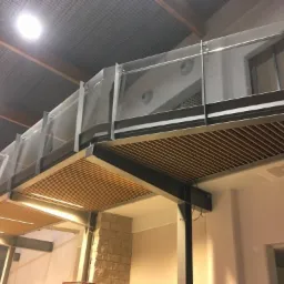 Nowoczesna antresola z drewnianym podbiciem i szklanymi balustradami, widok od dołu, częściowo widoczna drabina.