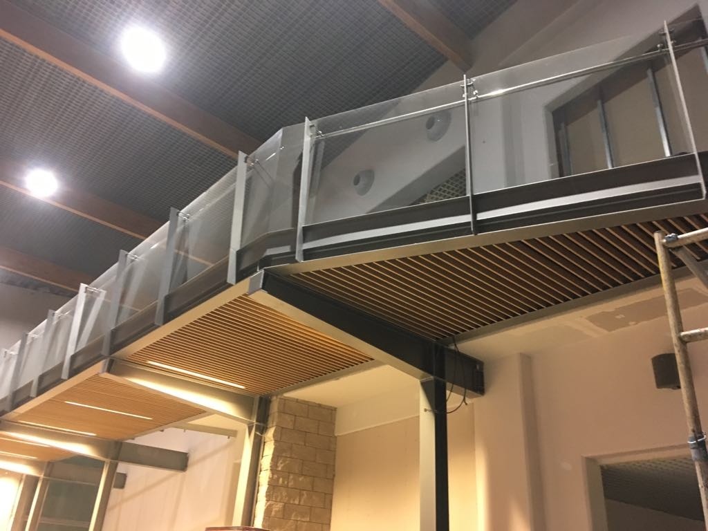 Nowoczesna antresola z drewnianym podbiciem i szklanymi balustradami, widok od dołu, częściowo widoczna drabina.