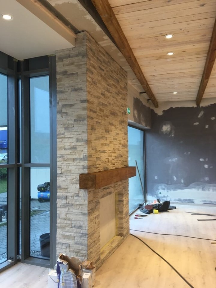 Wnętrze w trakcie wykończenia: obłożony kamieniem kominek z drewnianą półką, widoczne surowe drewniane belki na suficie, duże okna i elementy w trakcie prac wykończeniowych.