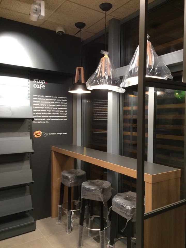Wnętrze stacji paliw Orlen w trakcie wykańczania: wysoki blat z drewnianymi nogami, trzy krzesła barowe owinięte folią ochronną, wiszące lampy w częściowo zdjętej folii, czarna ściana z logo 'stop...
