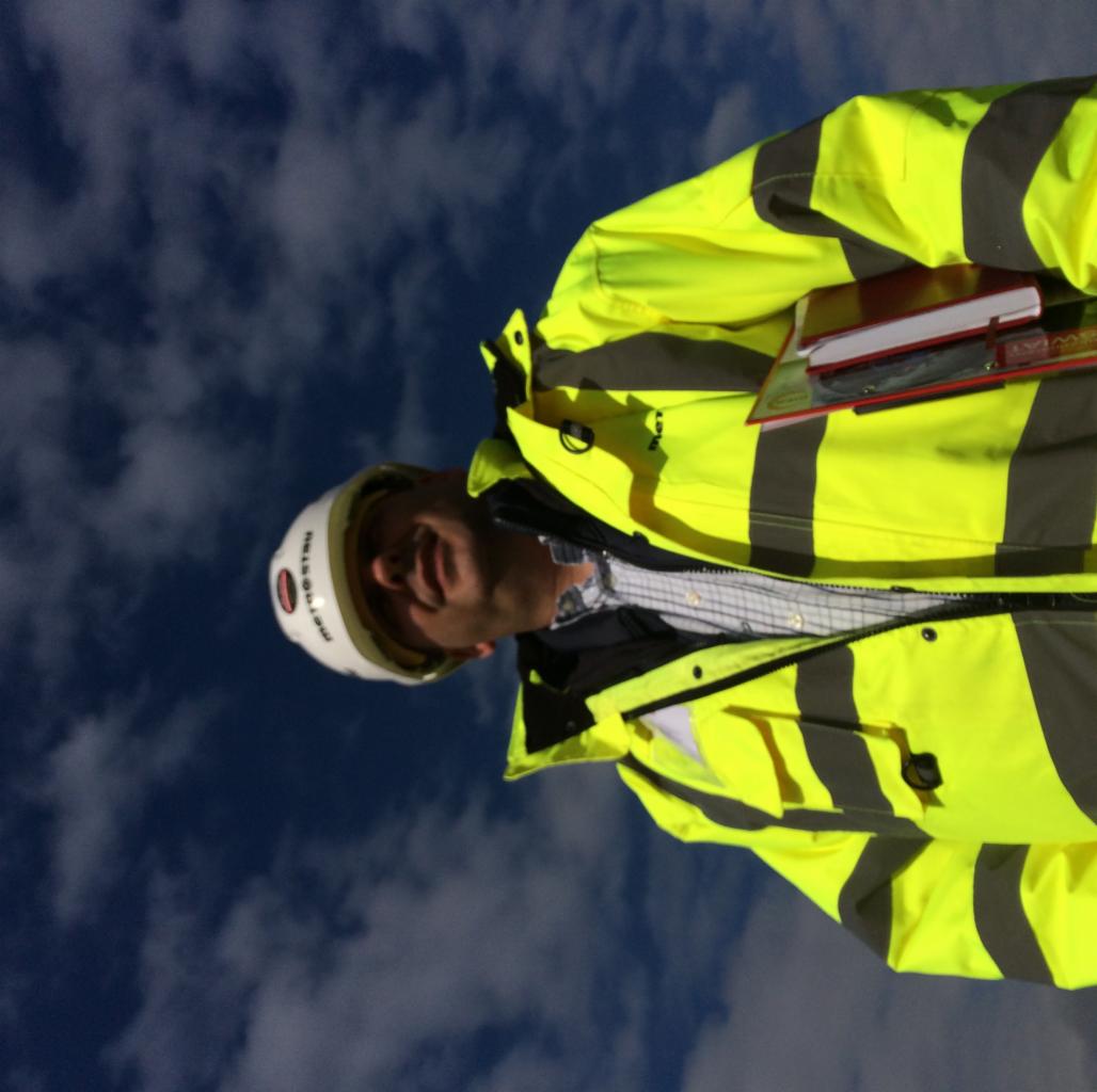 Mężczyzna w kasku i żółtej kurtce odblaskowej, trzymający notatnik, na tle błękitnego nieba z chmurami.