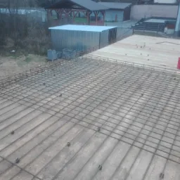 Zbrojenie płyty fundamentowej na drewnianym szalunku, widoczne czerwone i białe wiaderko oraz otoczenie placu budowy.