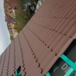 Układanie brązowej dachówki falistej na nowej konstrukcji z zielonych łat, widoczny fragment membrany dachowej z nadrukiem, w tle zabudowa mieszkalna.