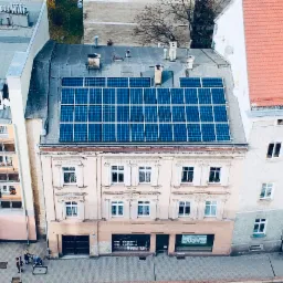 Panele słoneczne na dachu kamienicy w miejskiej zabudowie, widok z góry, błękitne moduły fotowoltaiczne równo ułożone na szarym dachu.