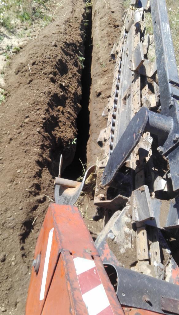 Wąski wykop w ziemi, widoczny mechanizm kopiący z pomarańczowym elementem z biało-czerwonymi pasami oraz łopata w tle.