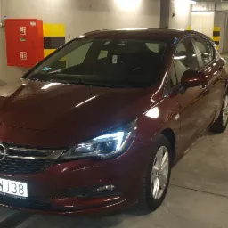 Czerwony samochód osobowy marki Opel Astra, zaparkowany w oświetlonym garażu podziemnym, widoczny z przodu i boku, z włączonymi światłami do jazdy dziennej.