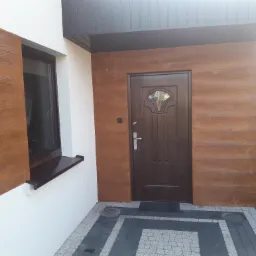 Elewacja domu z brązową okładziną imitującą drewno, brązowe drzwi wejściowe z szybą i szarą podbitką dachową, widoczne kostki brukowe na podłożu.