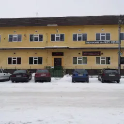 Dwupiętrowy budynek z napisem 'Producent odzieży męskiej' i 'Wyprzedaż' na fasadzie, z zaparkowanymi samochodami na śniegu przed budynkiem.