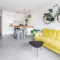 Nowoczesne wnętrze z szarą posadzką, żółtą sofą i rowerem na ścianie. Minimalistyczna kuchnia w tle. Jasne, przestronne pomieszczenie.