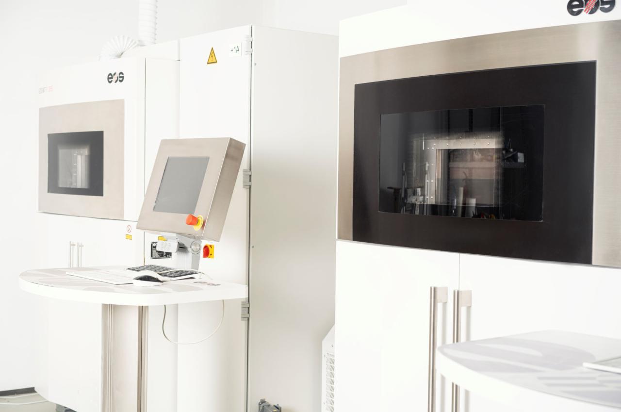 Dwa urządzenia do druku 3D z widocznymi komorami roboczymi, panel sterowania z klawiaturą i ekranem dotykowym, umieszczone w jasnym, sterylnym pomieszczeniu.