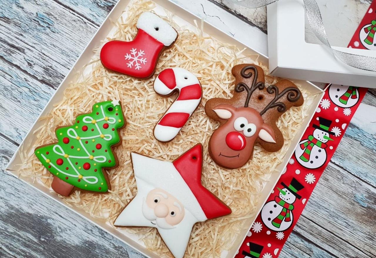 Świąteczne pierniczki w kształcie choinki, renifera, gwiazdy Mikołaja, skarpety i laski cukrowej ułożone w białym pudełku wypełnionym siankiem dekoracyjnym, obok czerwona wstążka z motywem bałwana.