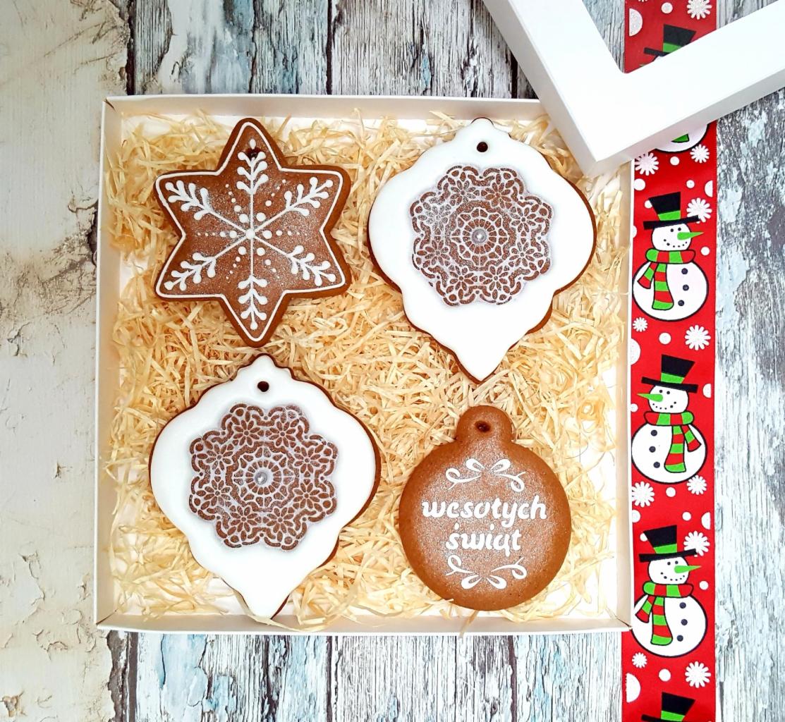 Świąteczny zestaw pierników dekoracyjnych w białym pudełku na słomkowym wypełniaczu, obok czerwona wstążka z motywem bałwanków.