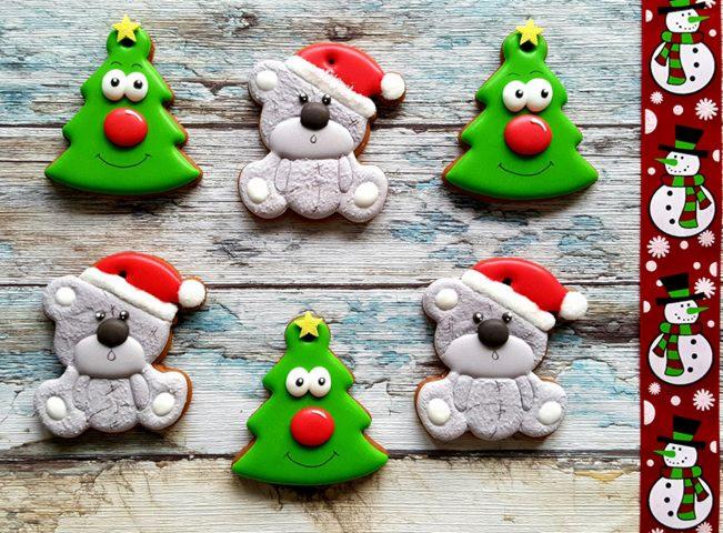 Świąteczne pierniczki: zielone choinki z czerwonymi nosami i uśmiechami, szare misie w czapkach Mikołaja, ułożone na drewnianym tle z dekoracyjną bordiurą ze śnieżynkami i bałwankami.