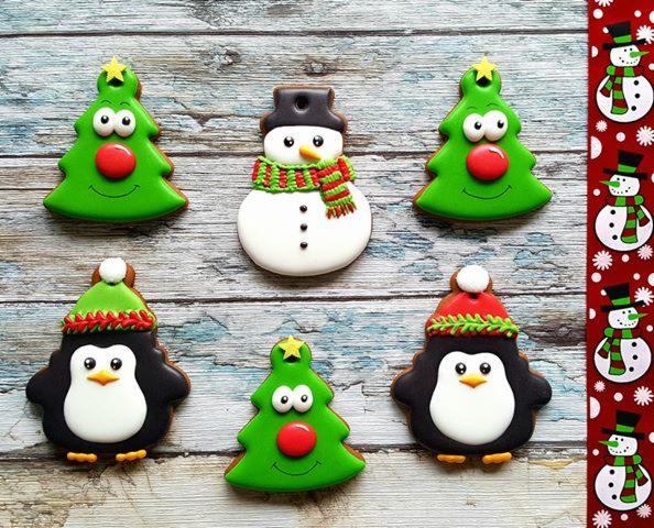 Świąteczne pierniczki dekoracyjne: choinki z oczami i czerwonym nosem, bałwanek w szaliku, pingwiny w czapkach, ułożone na drewnianym tle z motywem bałwanków.