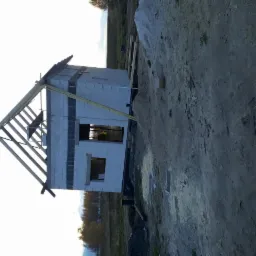 Surowy stan budowy domu jednorodzinnego z widoczną drewnianą konstrukcją dachu i pustakami, otoczonego ziemią i roślinnością.