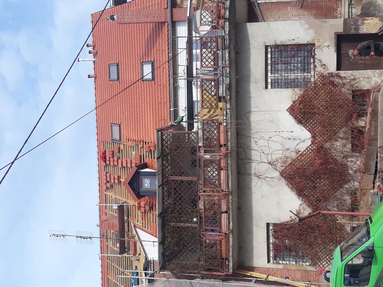 Częściowy widok dachu z czerwonej dachówki w trakcie układania, widoczne łaty i kontrłaty, okno dachowe oraz fragment elewacji sąsiedniego budynku z metalową kratą i pnącą roślinnością.