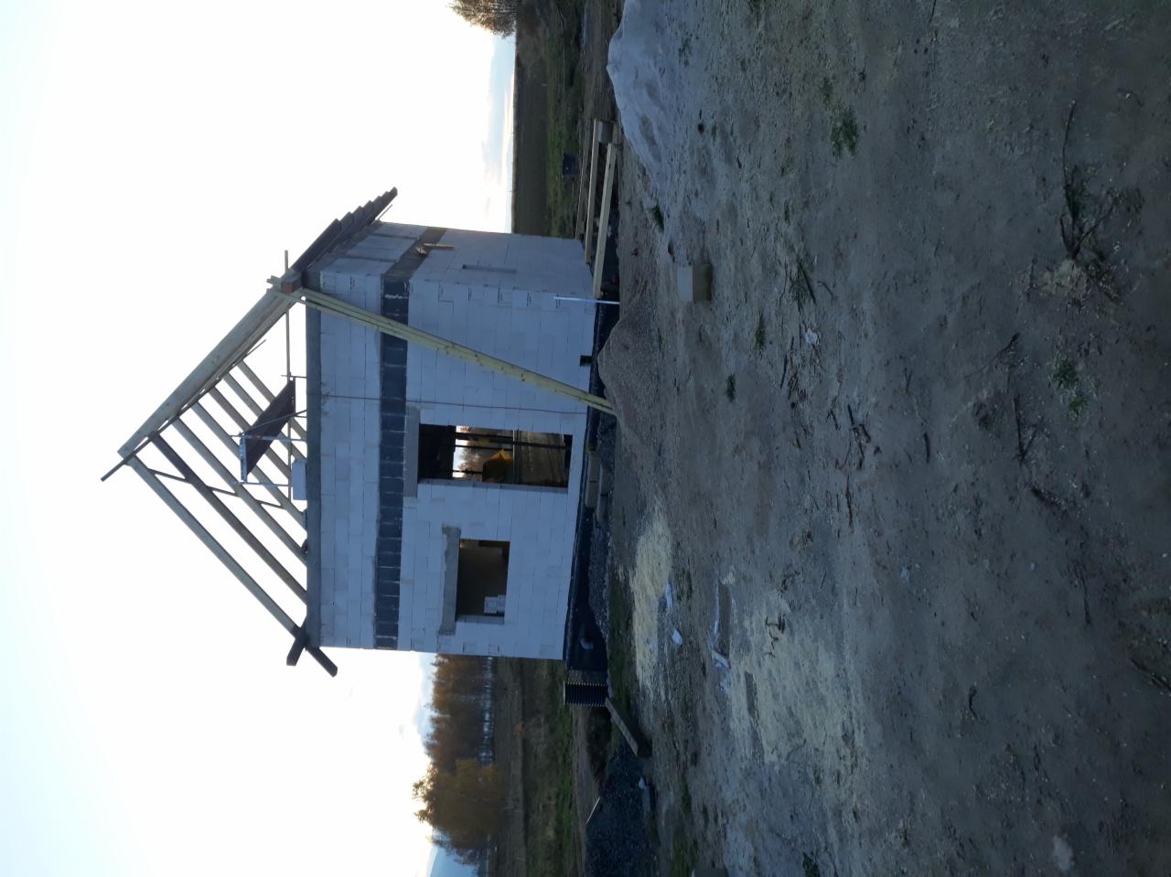 Surowy stan budowy domu jednorodzinnego z widoczną drewnianą konstrukcją dachu i pustakami, otoczonego ziemią i roślinnością.