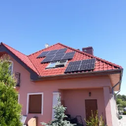 Dom jednorodzinny z czerwonym dachem, na kt&oacute;rym zamontowane są panele słoneczne; widoczny r&oacute;wnież satelita, okno dachowe i trawnik z kosiarkami.