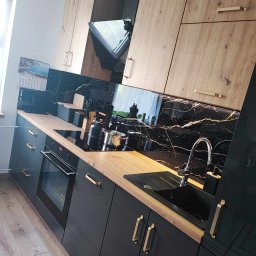 F.P.H.U. ROMEX - Nowoczesna kuchnia z czarnymi szafkami, drewnianymi blatami i marmurowym backsplash'em z złotymi żyłami. Widoczny czajnik na płycie indukcyjnej, zlew i bateria.