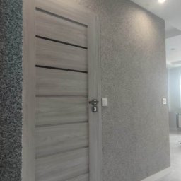 F.P.H.U. ROMEX - Jasnoszare drzwi z poziomymi wstawkami w wykończonym wnętrzu. Ściany pokryte dekoracyjnym tynkiem w odcieniach szarości. Biała, marmurowa podłoga i oświetlenie sufitowe.