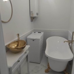 F.P.H.U. ROMEX - Biała łazienka z wolnostojącą wanną na złotych nóżkach, złotą umywalką na białej konsoli i białą pralką. Oświetlenie stanowią nowoczesne lampy wiszące.