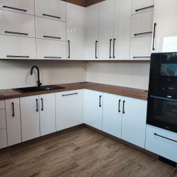 F.P.H.U. ROMEX - Biała kuchnia na wymiar z drewnianymi blatami i czarnymi uchwytami. Uwagę przykuwa czarny zlew i bateria. Podłoga wyłożona panelami imitującymi drewno.