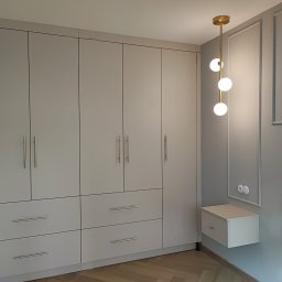 F.P.H.U. ROMEX - Jasnoszara szafa na wymiar z szufladami i złotymi uchwytami, uzupełniona o minimalistyczną lampę wiszącą i szafkę nocną, na tle ściany z dekoracyjnymi listwami.
