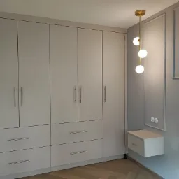 Jasnoszara szafa na wymiar z szufladami i złotymi uchwytami, uzupełniona o minimalistyczną lampę wiszącą i szafkę nocną, na tle ściany z dekoracyjnymi listwami.