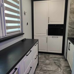 F.P.H.U. ROMEX - Nowoczesna kuchnia na wymiar z białymi frontami, czarnym blatem i zabudowaną piekarnikiem. Widoczne żaluzje okienne i marmurowe płytki podłogowe.