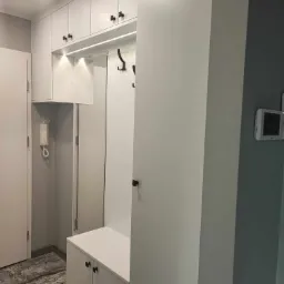 Biała szafa wnękowa z siedziskiem i wieszakami w przedpokoju. Szafki górne z oświetleniem LED. Ciemne uchwyty szafek. Na ścianie domofon.