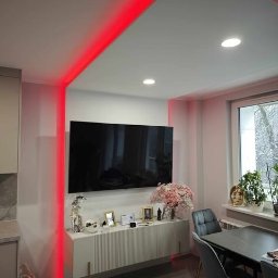 Firma Produkcyjno-Handlowo-Usługowa ROMEX Alicja Niezgoda - Nowoczesny salon z podświetleniem LED w kolorze czerwonym, szafka RTV z dekoracjami, telewizor na ścianie i stół z krzesłami. Minimalistyczny design wnętrza.